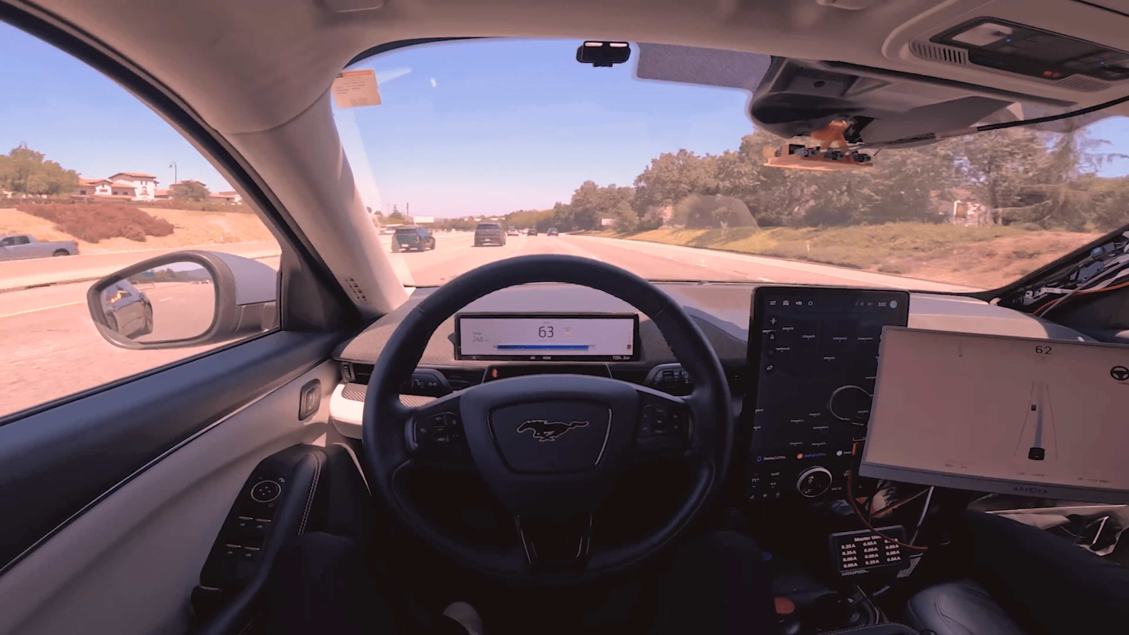 Interior of the IDrive demonstration vehicle on a freeway, showing the L2+ driver assistance camera module behind the rear-view mirror and the real-time driver display.