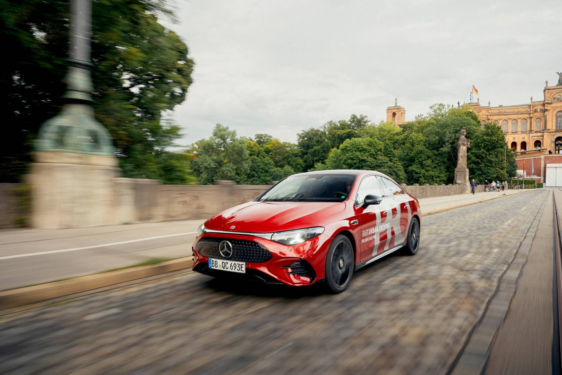 Red Mercedes-Benz in PRO livery, illustrating MB.DRIVE ASSIST PRO, driving on a cobbled city street past historic buildings